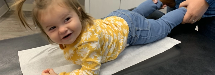 A child lying face down on a treatment table covered with paper, while a practitioner gently holds the child’s legs during an adjustment.