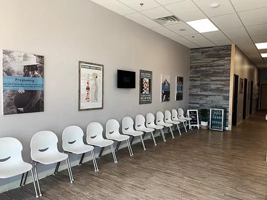 A spacious waiting area featuring a row of white chairs along a light gray wall, decorated with health-related posters and a mounted screen. The flooring is wood-textured, and the space has bright overhead lighting.