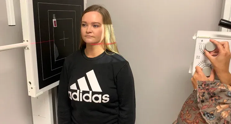 A patient positioned for an X-ray scan in front of a black panel with laser alignment lines, while the practitioner prepares the equipment.