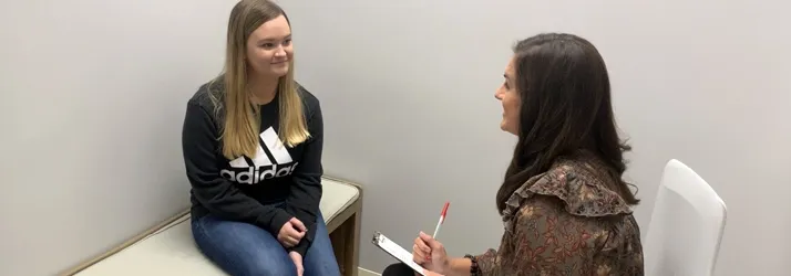 A patient seated on a bench while Dr. Rachel Franco takes notes on a clipboard during a private consultation in a neutral-toned room