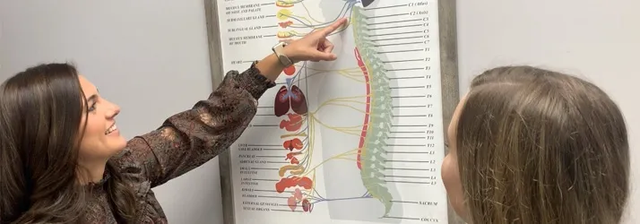 Dr. Rachel Franco pointing to a detailed anatomical chart of the spine and nervous system during an educational discussion with a patient in a consultation room.