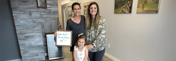Dr. Rachel Franco, a woman and a child standing in a clinic hallway, holding a sign with handwritten feedback. The background includes gray textured walls and framed wellness posters.