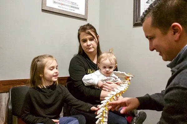 An adult seated with two children in a consultation room while Dr. Tony Franco holds a spinal model, explaining chiropractic concepts. Background features framed educational posters.