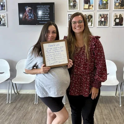 Dr. Rachel Franco and patient standing in a waiting area, one holding a sign with handwritten feedback. The background features white chairs and a wall decorated with framed photos.