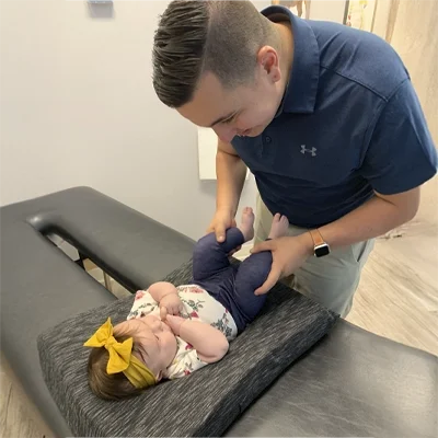 An infant lying on a padded surface while Dr. Tony Franco gently adjusts the legs. The setting includes a treatment table and neutral-colored walls.